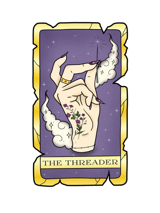 A weathered tarot card featuring an elegant hand, with long purple nails, a gold jewelled ring, and a flower tattoo, holding a sparkling embroidery needle. The tarot card reads "The Threader".The hand is surrounded by tattoo style clouds with stars on a purple background.