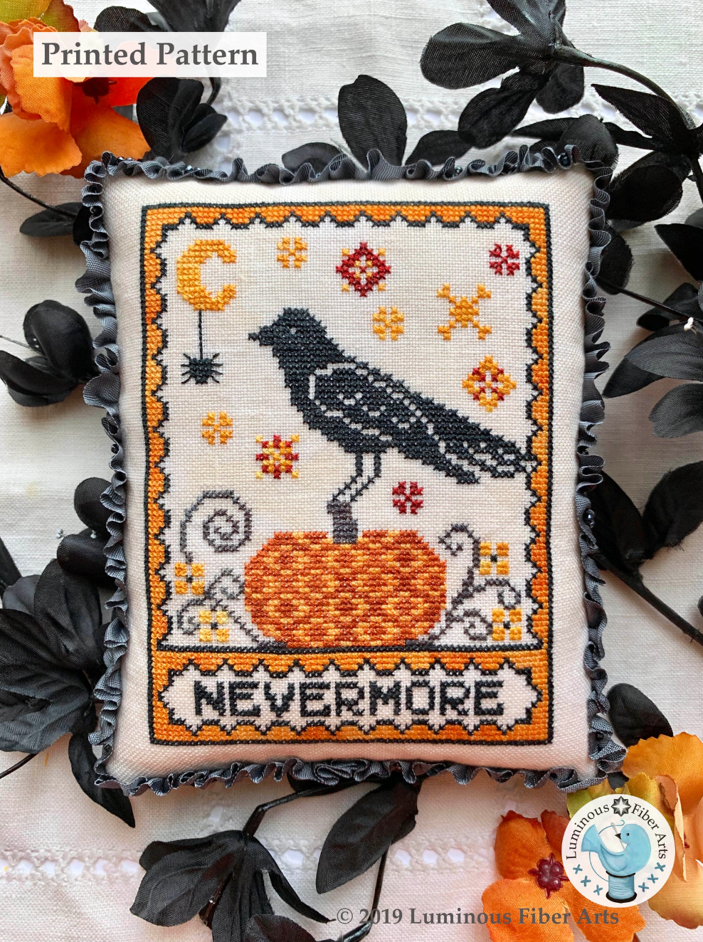 A cross stitch piece finished as a small pillow features a raven sitting atop a pumpkin surrounded by stars and a moon with a spider hanging off it